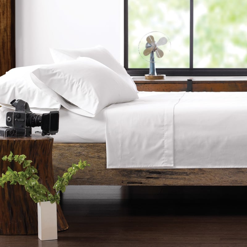 A neatly made bed with WestPoint Home’s Martex Green 250 white sheets and pillowcase sits beside a rustic wooden nightstand holding a vintage camera, a vase with a plant, and near a window overlooking greenery and a small fan.
