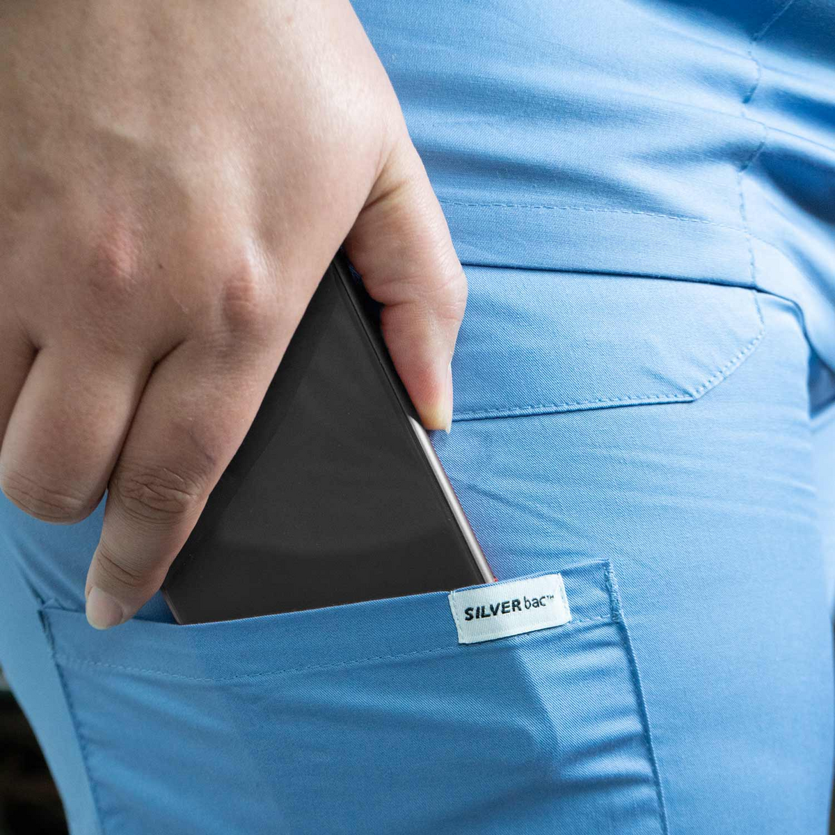 A hand slips a black phone into the back pocket of light blue pants with a visible SILVERbac label, highlighting WestPoint Home’s Martex Health Classic Scrub Tops—antimicrobial scrubs for everyday protection.