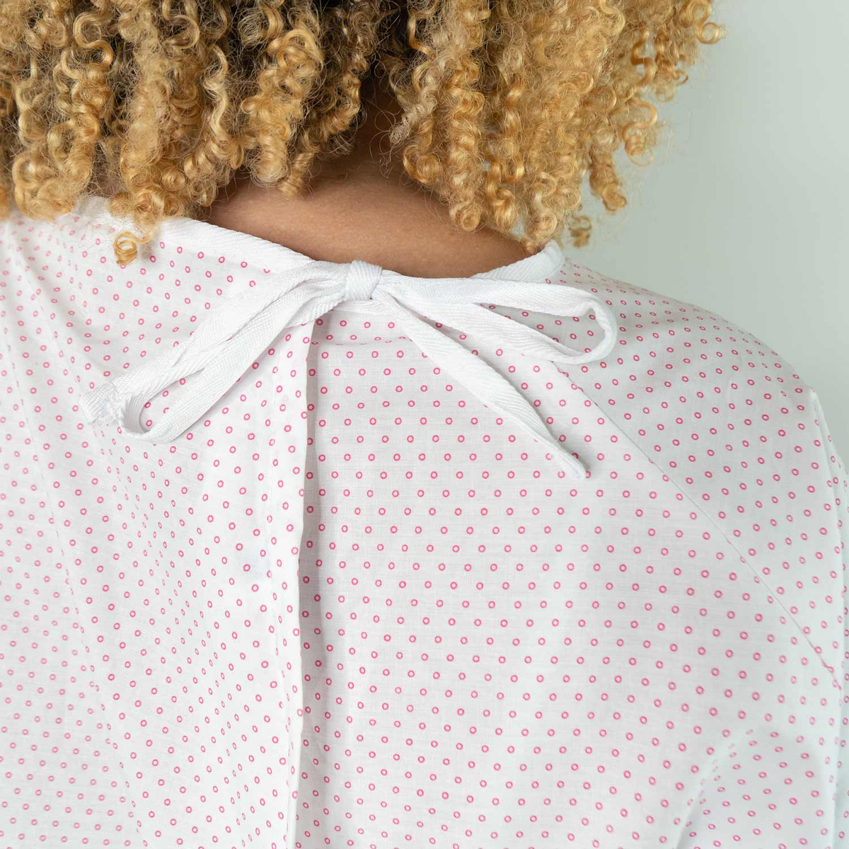 Close-up of a person with curly hair wearing the Martex Health Lab Coat by WestPoint Home, featuring small pink dots and a back tie. The image highlights upper back comfort and modern style in this patient gown.
