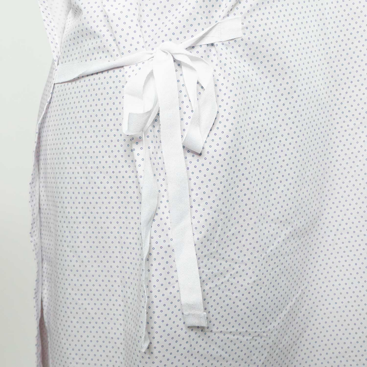 A close-up of a person wearing Martex Health Lab Coats by WestPoint Home, featuring white fabric with small blue dots, tied at the waist, and enhanced with SILVERbac antimicrobial technology for added protection.