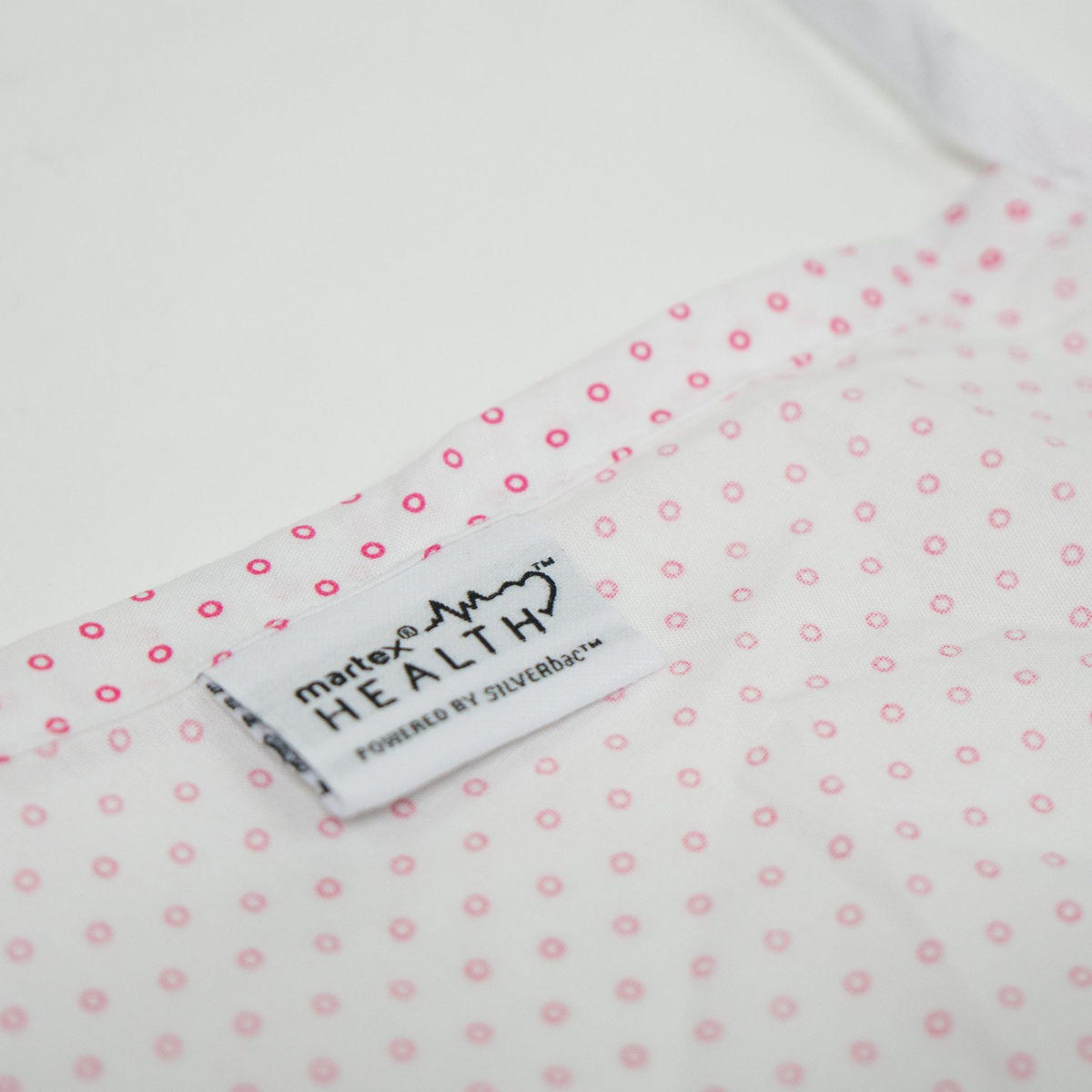 Close-up of a white fabric with small pink dots and a label reading matrix HEALTH™ POWERED BY SILVERbac™, from Martex Health Lab Coats by WestPoint Home. This textile offers advanced antimicrobial technology for enhanced protection.