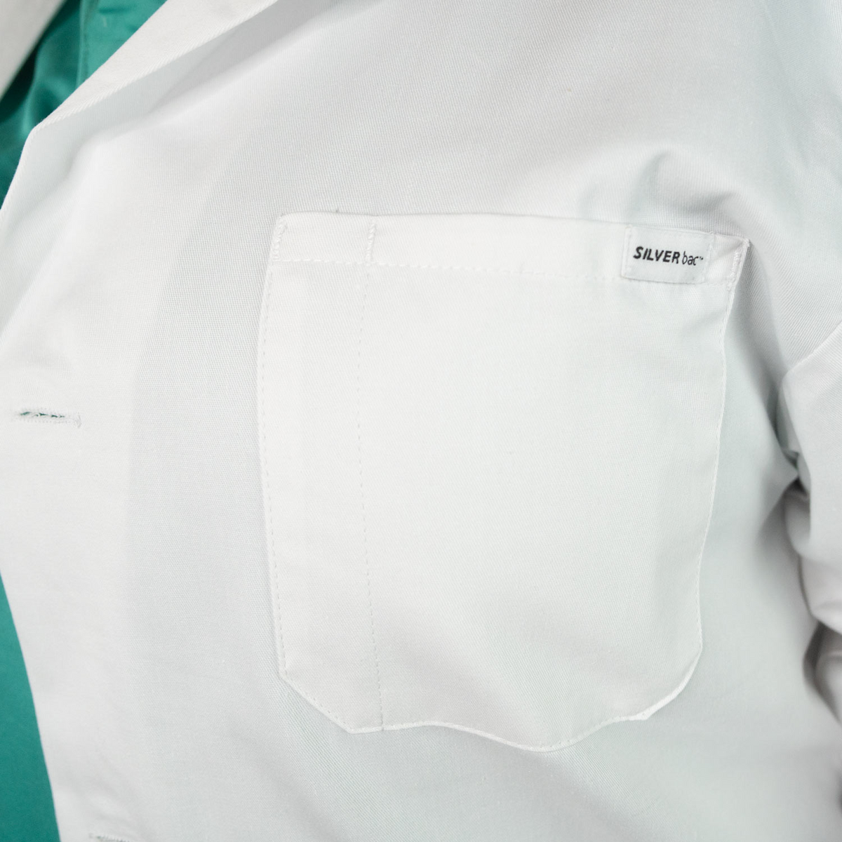 Close-up of the chest pocket on a white Martex Health Lab Coat by WestPoint Home, with a visible Silver tag above the pocket and a green shirt underneath, highlighting innovative SILVERbac technology.