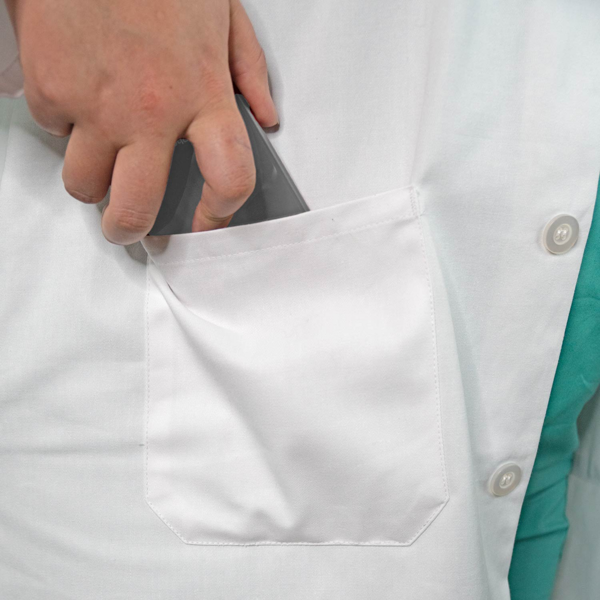 A person wearing a Martex Health Lab Coat by WestPoint Home with SILVERbac technology is placing a smartphone into its front pocket. Only the hand, part of the phone, and the coat are visible.