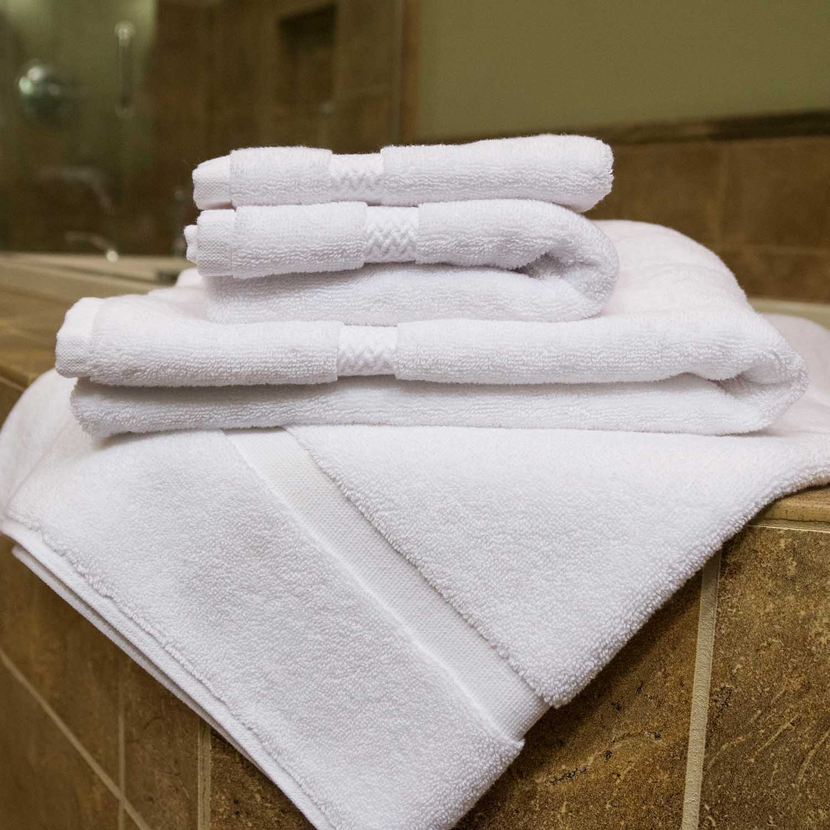 A stack of neatly folded white Martex Health Towels by WestPoint Home rests on the edge of a brown tiled bathtub in a bathroom.