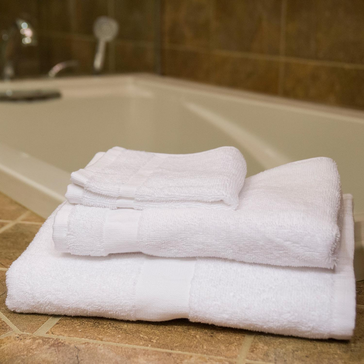 A neatly folded stack of Martex Health Towels from WestPoint Home rests on a tiled surface beside a bathtub in a bathroom with brown tile walls.