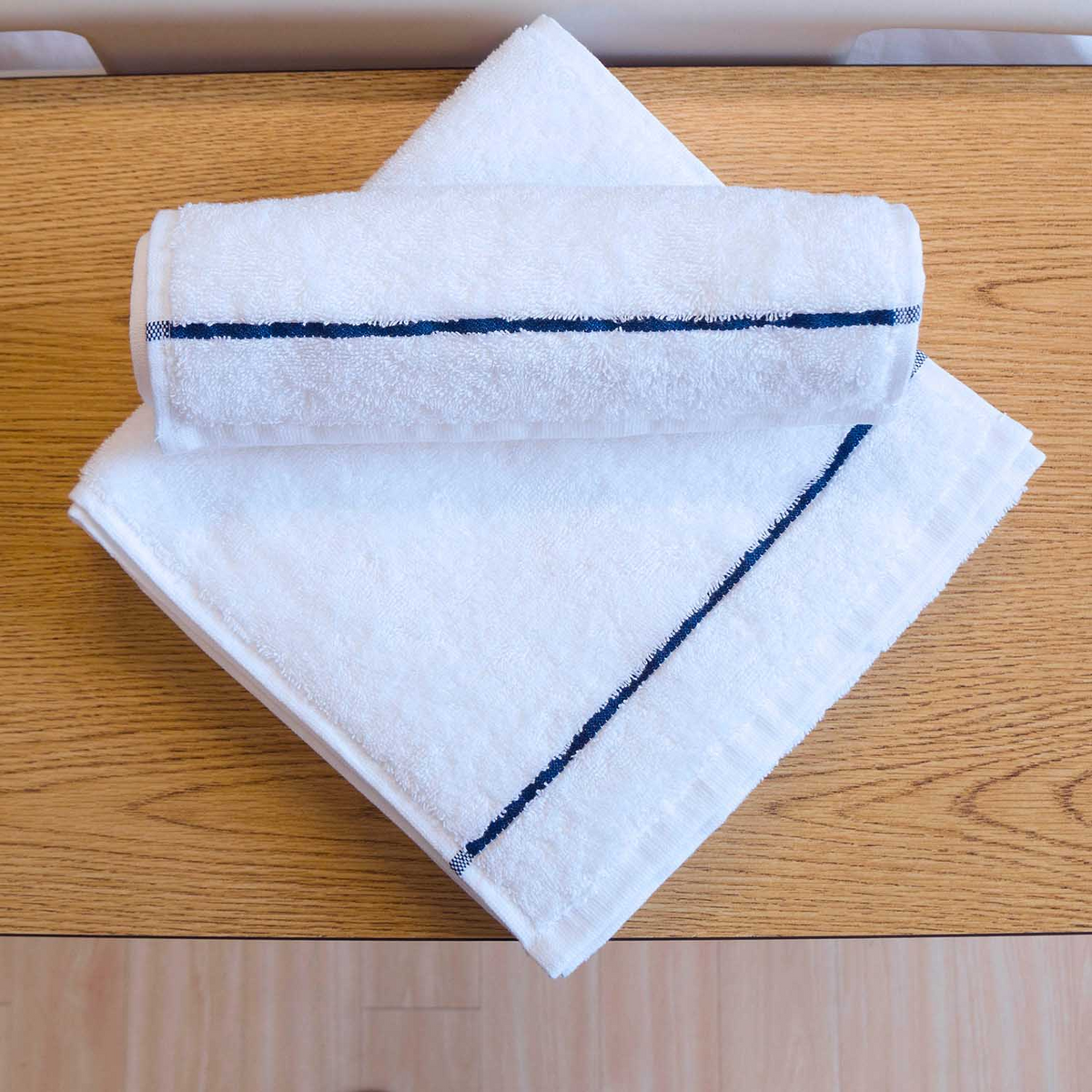 A neatly folded Martex Active Towel by WestPoint Home, featuring a dark blue edge stripe, rests on a wooden surface, with one towel rolled and placed atop another antibacterial towel.