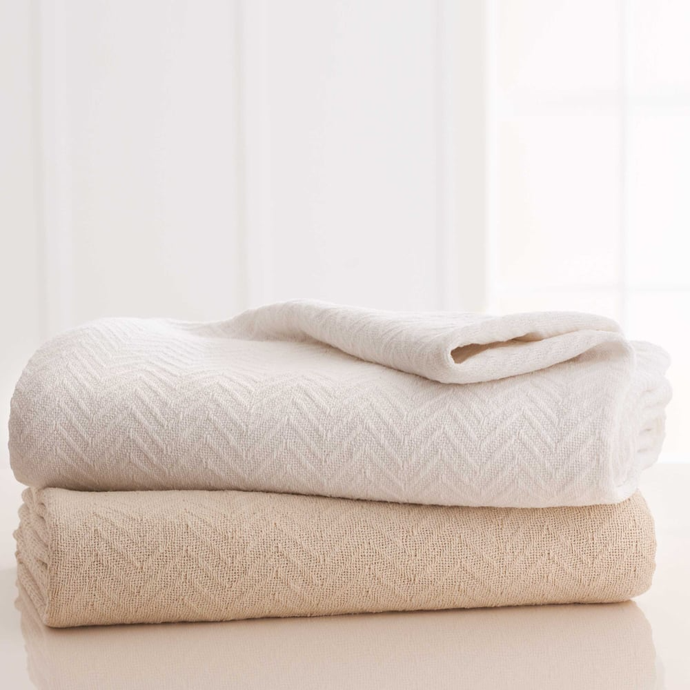 Two Grand Patrician Cotton Blankets by WestPoint Home, in cream and beige, are stacked on a light surface with a soft background. Subtle chevron texture makes them ideal for hotel use or bulk wholesale blanket orders.