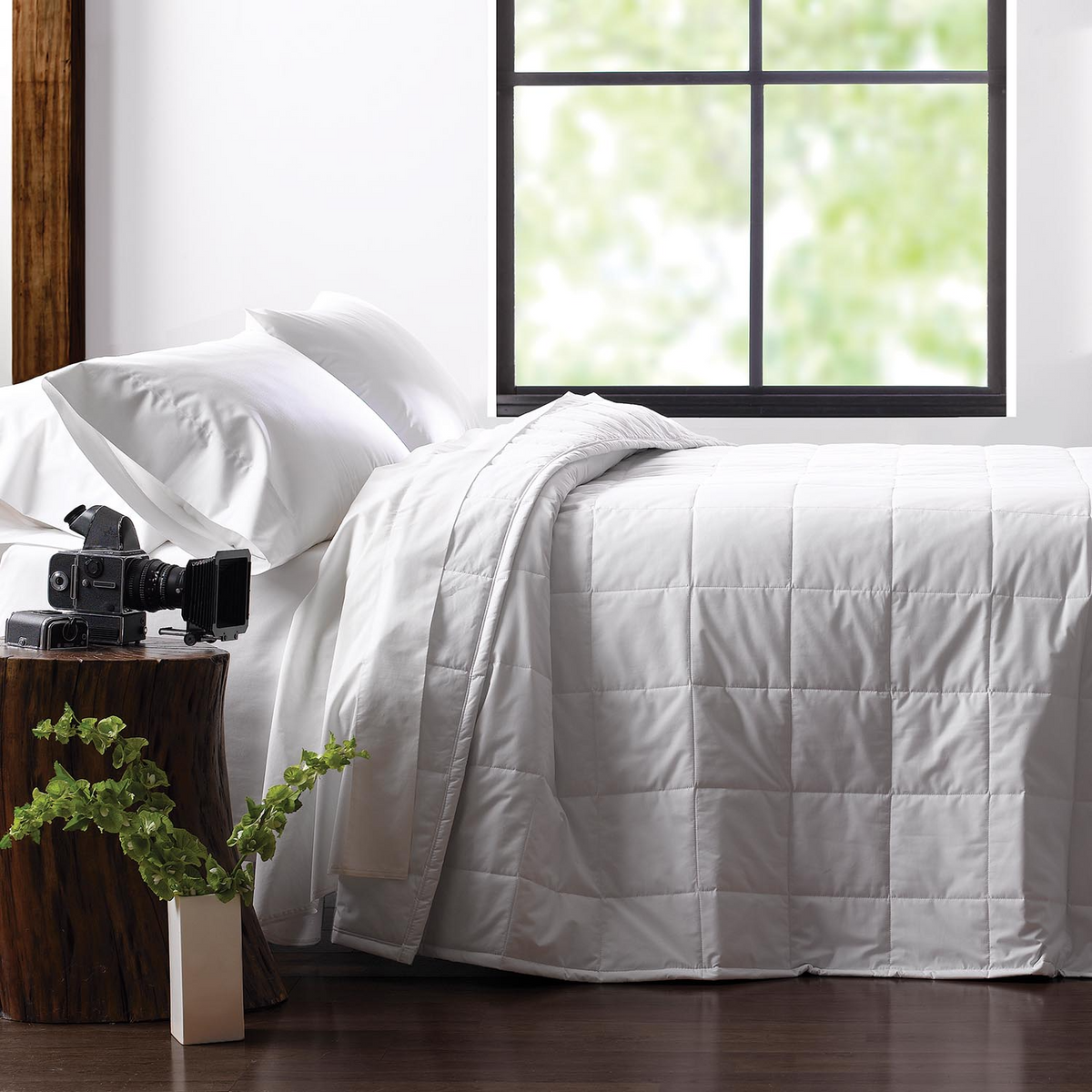 A neatly made bed with white bedding features the Martex Basics - Down Alt Quilted Blanket by WestPoint Home, filled with EcoPure polyester fiber. Nearby, a vintage camera rests on a wooden stump nightstand next to a small white vase.