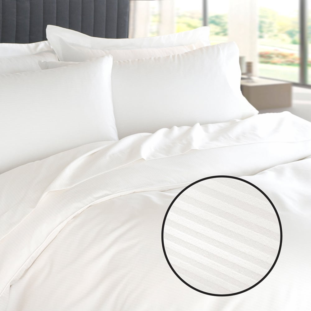 A neatly made bed with the Martex Basics - Down Alt Quilted Comforter by WestPoint Home features white striped bedding and pillows. An inset shows a close-up of the subtle stripe pattern, as natural light streams in through a window behind.