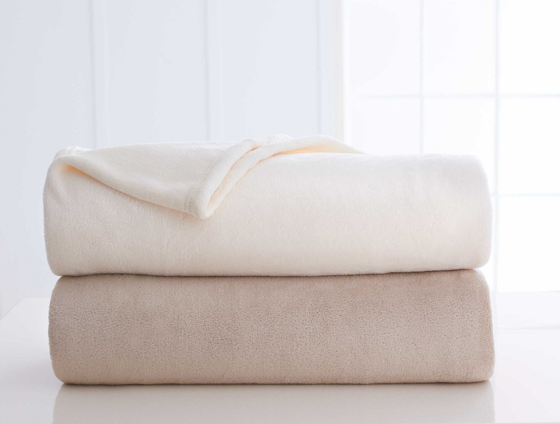 Two plush Martex blankets by WestPoint Home are stacked on a white surface. The top is cream, the bottom is light beige. A softly lit window and white walls in the background create a calm, cozy atmosphere.