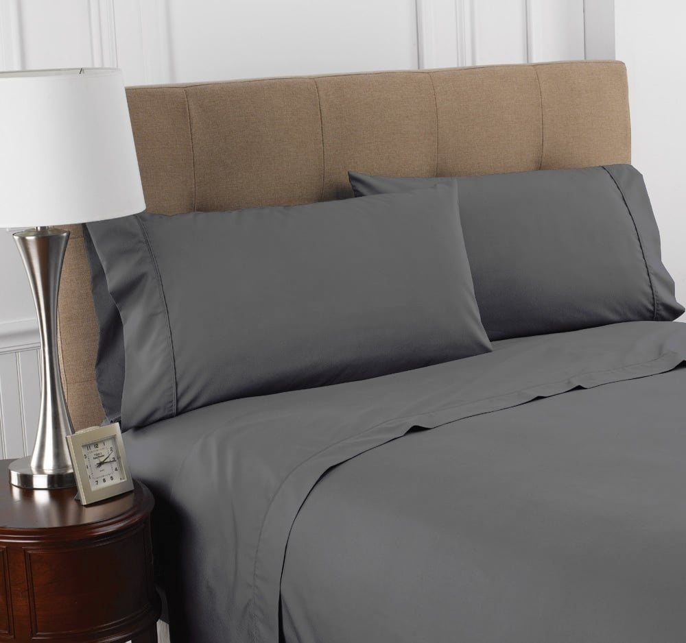 A neatly made bed featuring WestPoint Homes Martex Colors 200 pillow cases in deep gray, a beige upholstered headboard, a round wooden nightstand with a silver lamp, and a small picture frame.