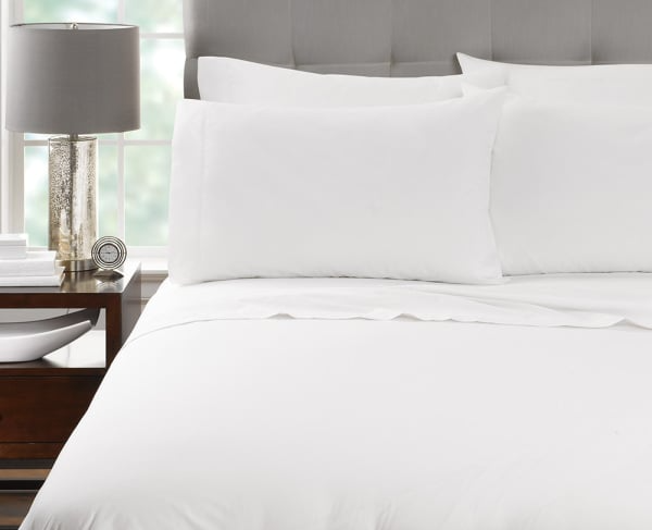 A neatly made bed featuring WestPoint Home Millennium - Duvet Covers and white pillows stands by a wooden nightstand with a modern lamp, small dish, and decorative clock in a bright room with a large window.