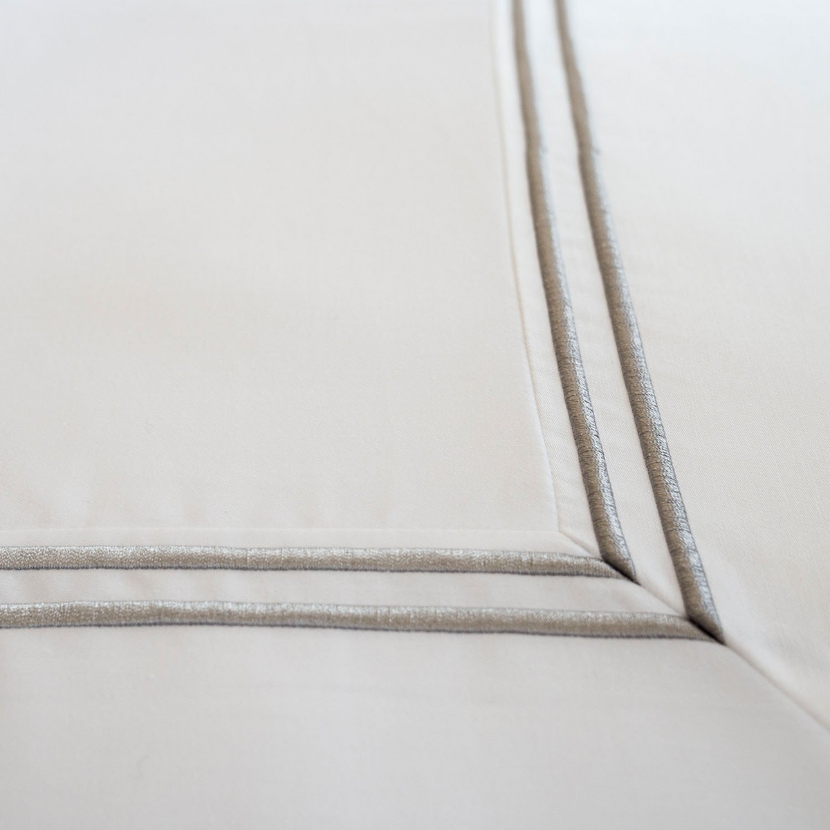 Close-up of the Gryphon Collection - Duvet Cover - Solid by WestPoint Home in beige Supima cotton, featuring two parallel metallic silver embroidered lines at the corner. The fabric is smooth and neatly stitched, evoking luxury hotel bedding.