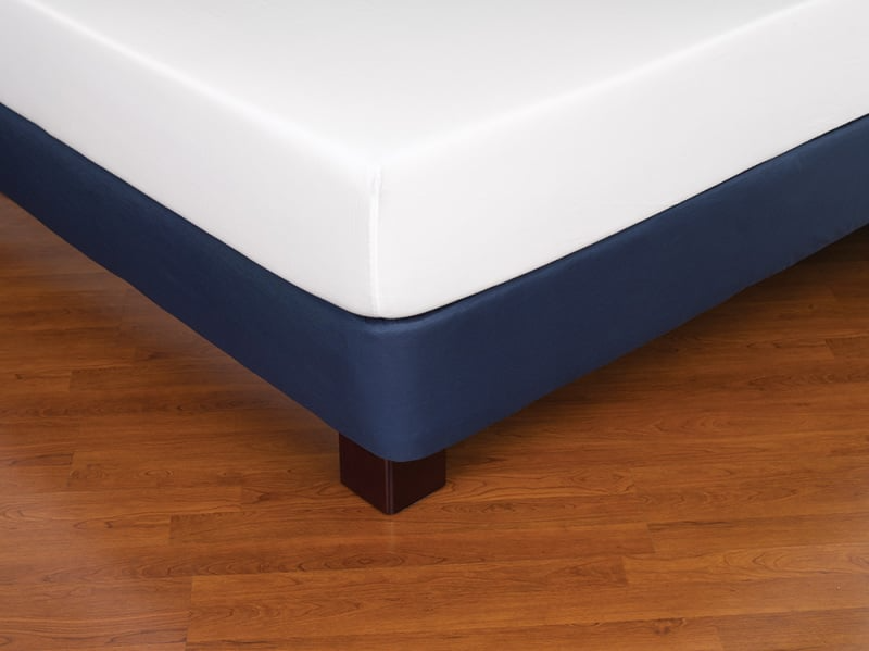 A close-up of a neatly made bed with a white mattress and navy Bedskirts - Westpoint Hospitality by WestPoint Home, featuring EcoPure polyester fiberfill, on dark wooden legs atop a polished wooden floor.
