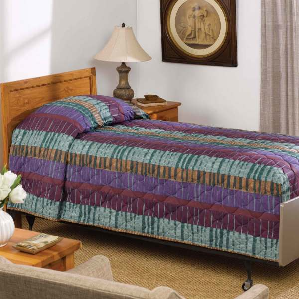 A twin bed features the WestPoint Home Martex Mainspreads Pattern bedspread, a colorful striped quilt filled with EcoPure polyester fiberfill for comfort, complemented by a wooden headboard, lamp-lit nightstand, framed picture, and beige armchair.