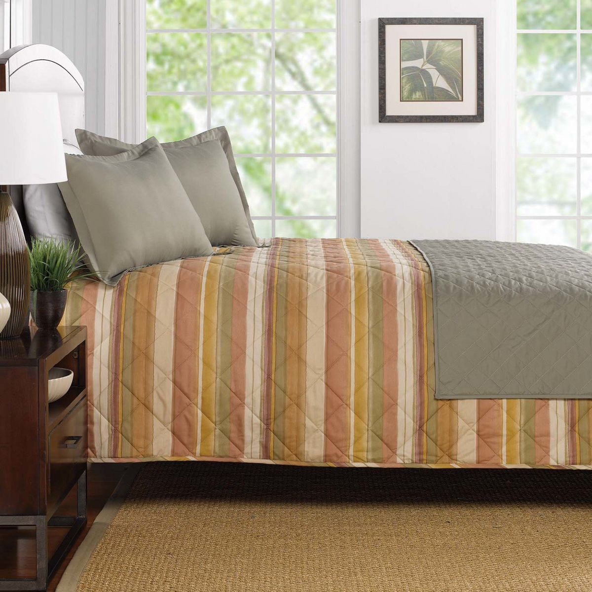 A bed featuring WestPoint Homes Bedskirts - Westpoint Hospitality - Pattern (Copy), with green and tan striped bedding, matching pillows, a quilted green EcoPure polyester blanket, large windows, a leaf wall print, and a nightstand with lamp and plant.