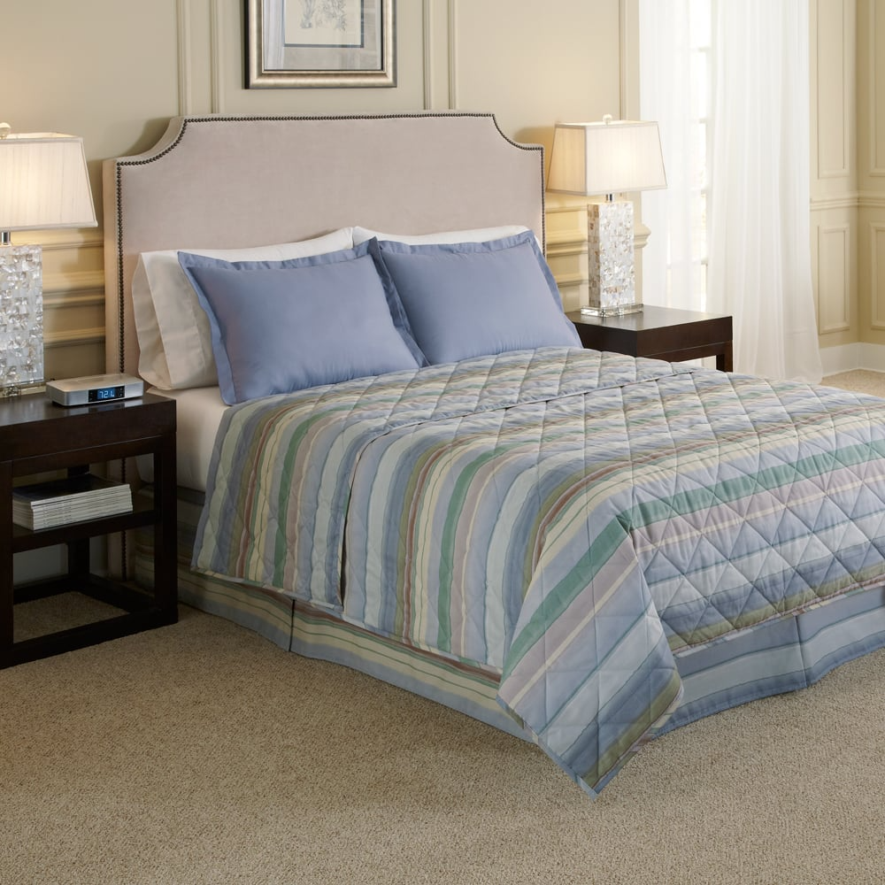 A neatly made bed featuring a WestPoint Home Bedskirt from the Westpoint Hospitality collection, accented by blue pillows and dark wood nightstands with crystal lamps, in an elegant bedroom with beige walls and carpet.