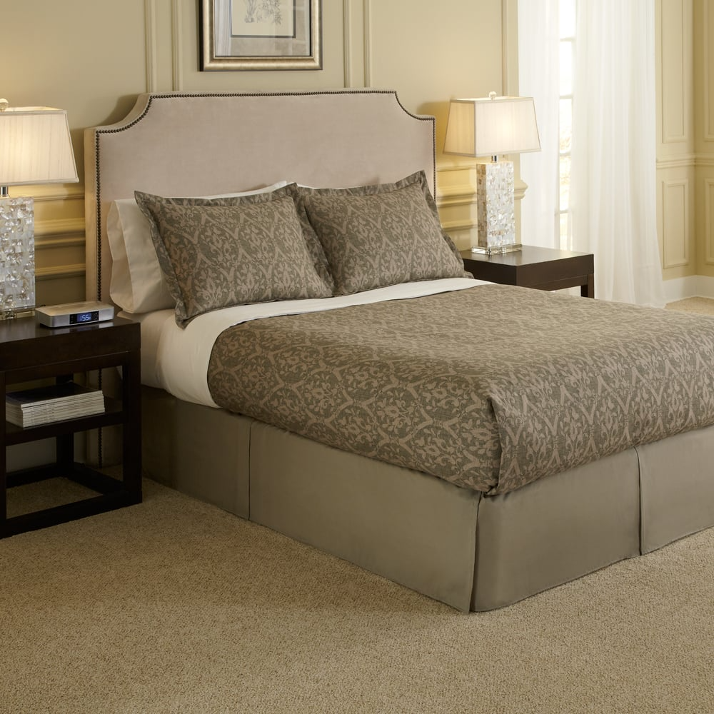 A neatly made bed features WestPoint Home Bedspreads - Solids with EcoPure polyester fiberfill and two matching pillows, set between dark wood nightstands with lamps in a softly lit, beige-toned, carpeted bedroom.