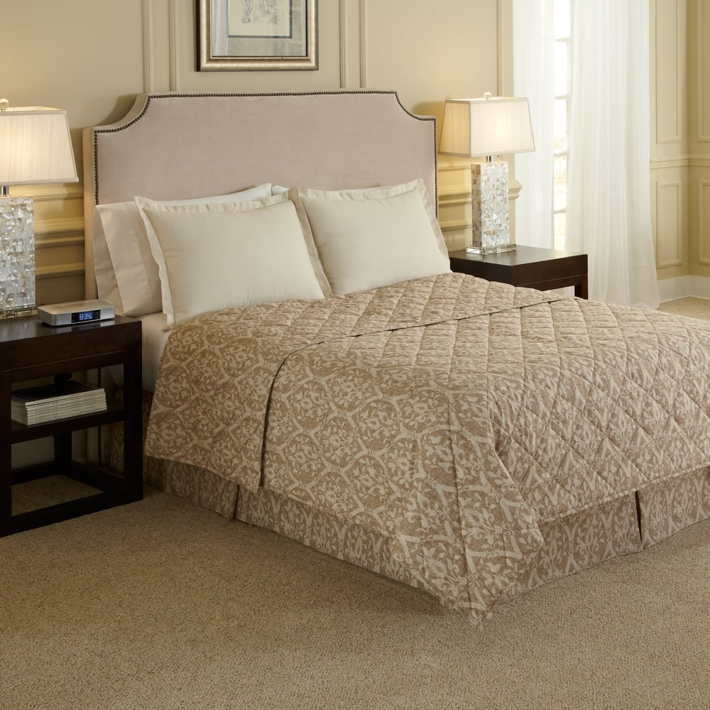 A neatly made bed featuring a WestPoint Home bedskirt from the Westpoint Hospitality Pattern collection, accented with three cream pillows, stands between dark nightstands in an elegant bedroom with softly lit tan walls and carpet.