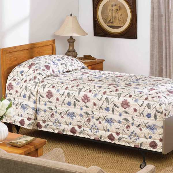 A neatly made single bed features the WestPoint Home Bedspread - Martex Mainspreads - Pattern, and sits next to a wooden nightstand with a lamp in a cozy room adorned with a framed picture and neutral curtains.