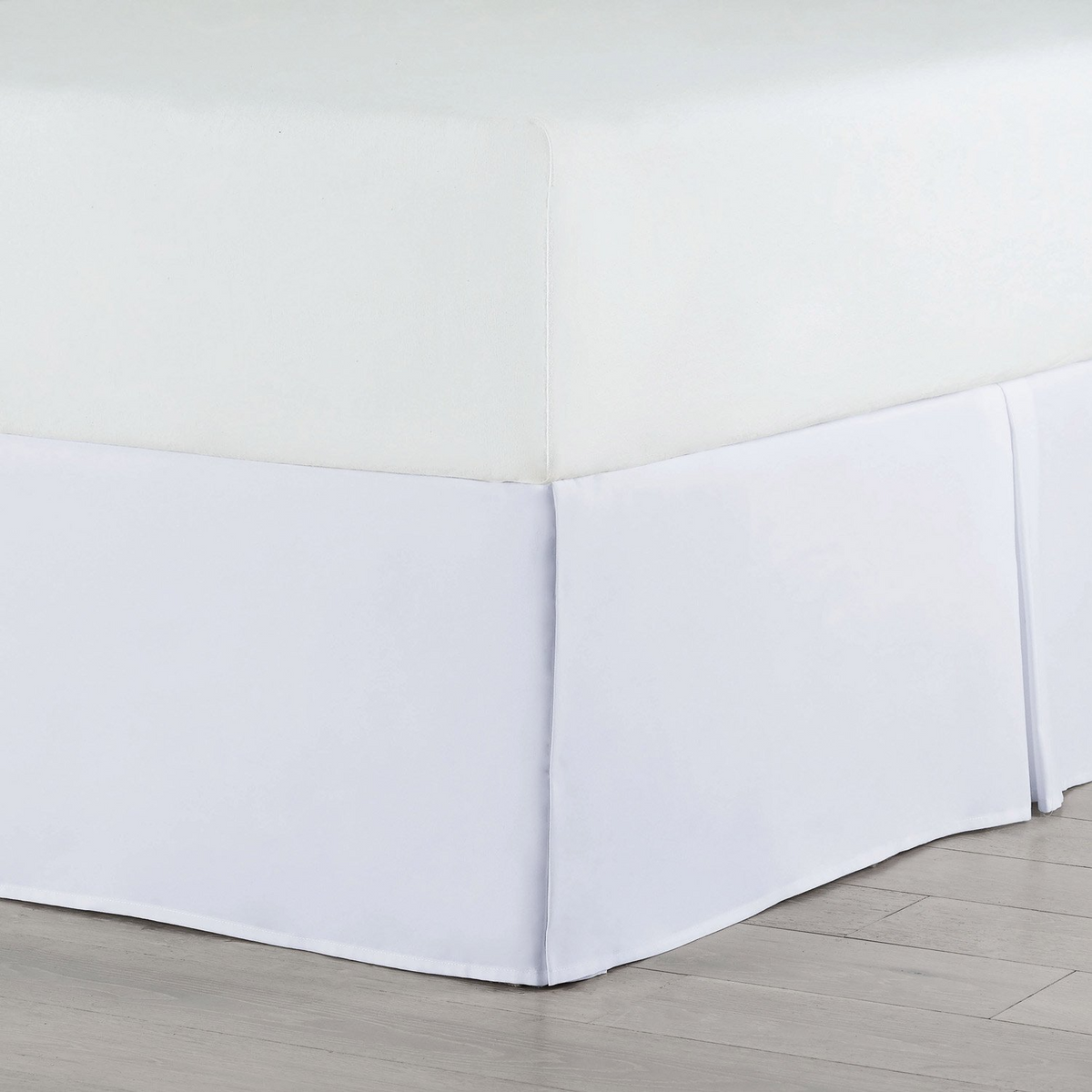 A close-up of a bed corner shows the Martex Basics Bedskirt by WestPoint Home in crisp white, neatly draping to the floor over a plain mattress, resembling hotel bedding and placed on a light wood floor.