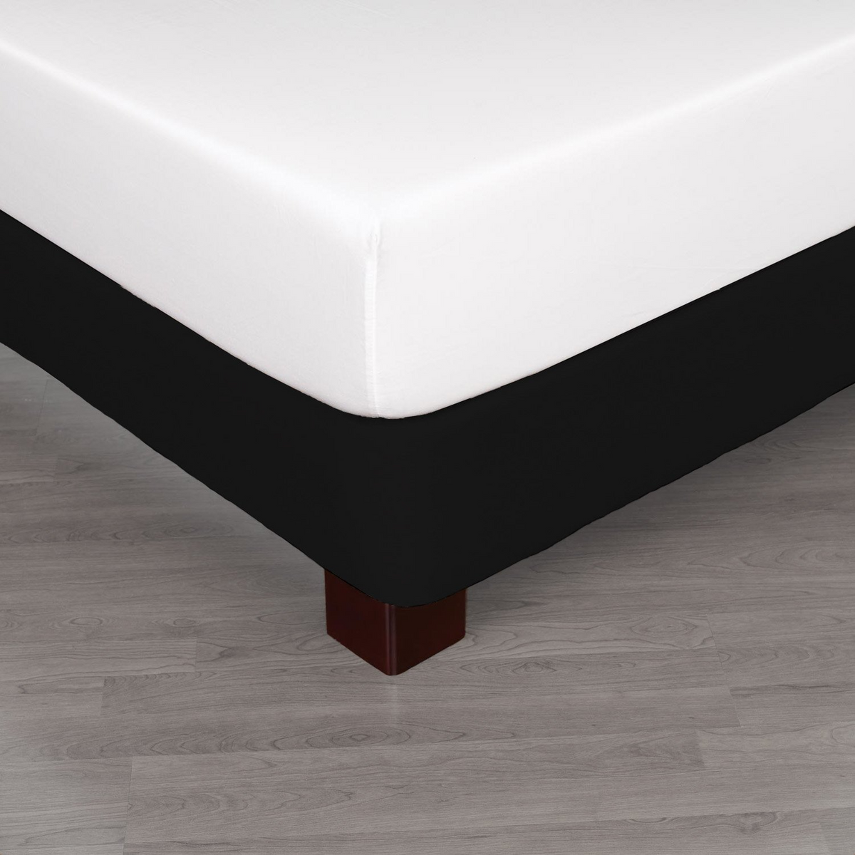 A close-up of a bed corner shows crisp hotel bedding with a Martex Basics - Bedskirts by WestPoint Home in black, a white mattress, and wooden legs on a light hardwood floor.