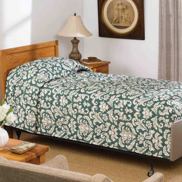 A neatly made single bed features a green and white floral-patterned Martex Mainspreads bedspread by WestPoint Home, GSA compliant, next to a wooden nightstand with a lamp and phone in a cozy, carpeted bedroom with framed artwork.
