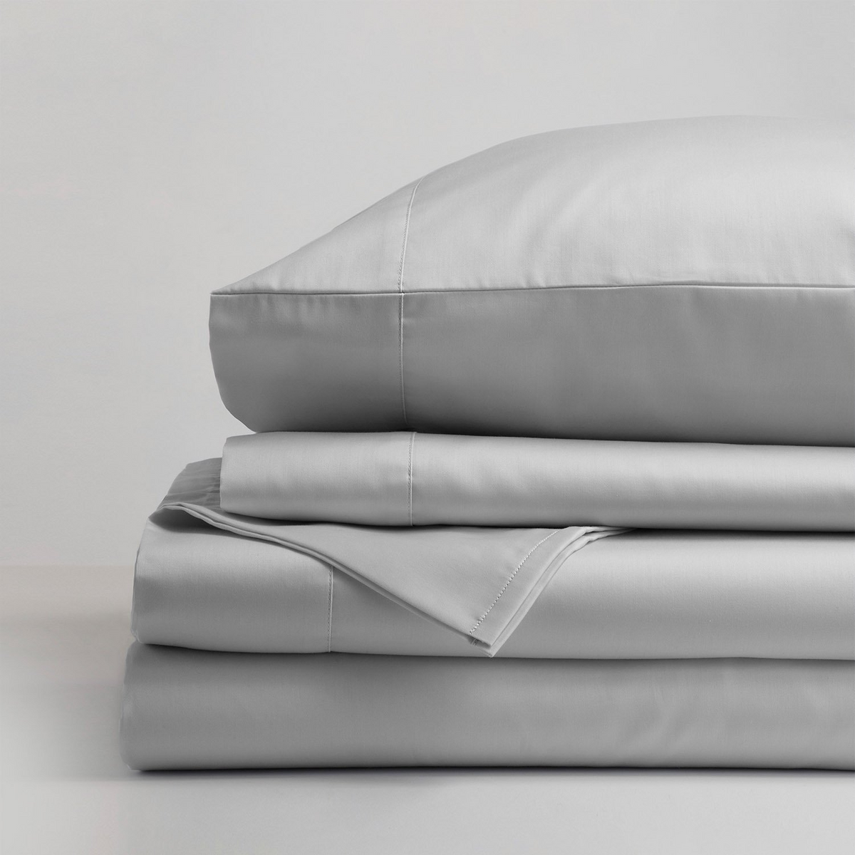 A neatly folded stack of Gryphon Collection - Sheets - Solid by WestPoint Home sits on a white surface, topped with a pillow, all showcasing smooth and clean luxury fabric.