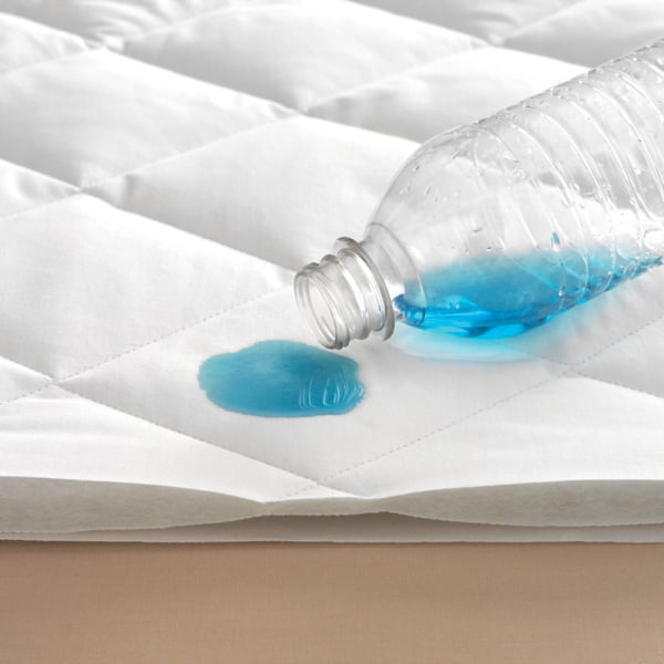 A clear plastic bottle pours blue liquid onto the Martex Ultra Touch Mattress Pad by WestPoint Home, showcasing the pad’s excellent water-resistant properties.