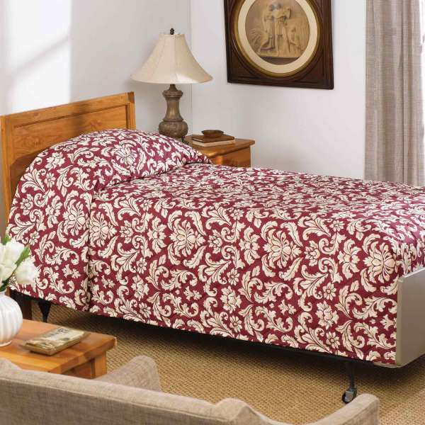 A neatly made single bed with a red and white floral-patterned WestPoint Home Martex Mainspreads bedspread, GSA compliant, sits beside a wooden nightstand with a lamp and framed picture. Beige carpet and light curtains finish the room.