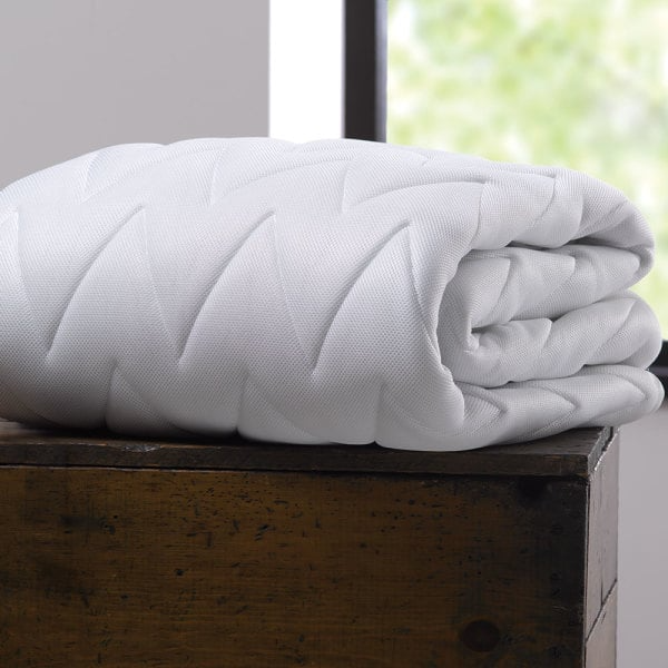 The WestPoint Home Mattress Pad - Martex Ultra Touch, a white quilted waterproof pad with a zigzag pattern and EcoPure fiberfill, is folded on a wooden surface near a sunlit window.
