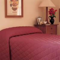 A neatly made bed with a maroon WestPoint Home Bedspread - Martex Mainspreads - Solid sits beside a wooden nightstand holding a lamp, red flowers in a vase, framed photos, and a cup, with artwork hanging above.