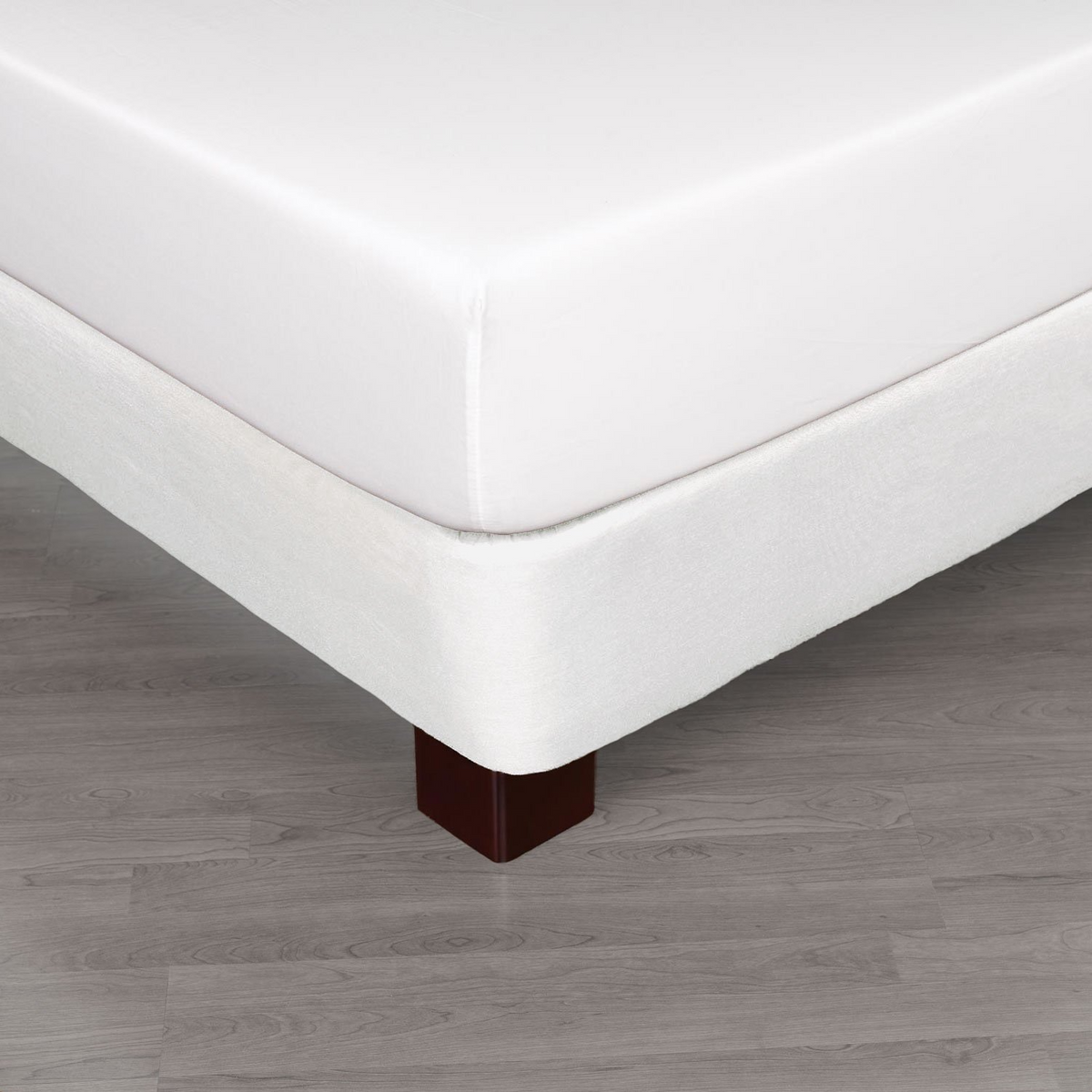 A close-up of a bed corner with crisp hotel bedding, showcasing a white Martex Basics - Bedskirts by WestPoint Home over a matching base with dark wooden legs on a light gray wooden floor.
