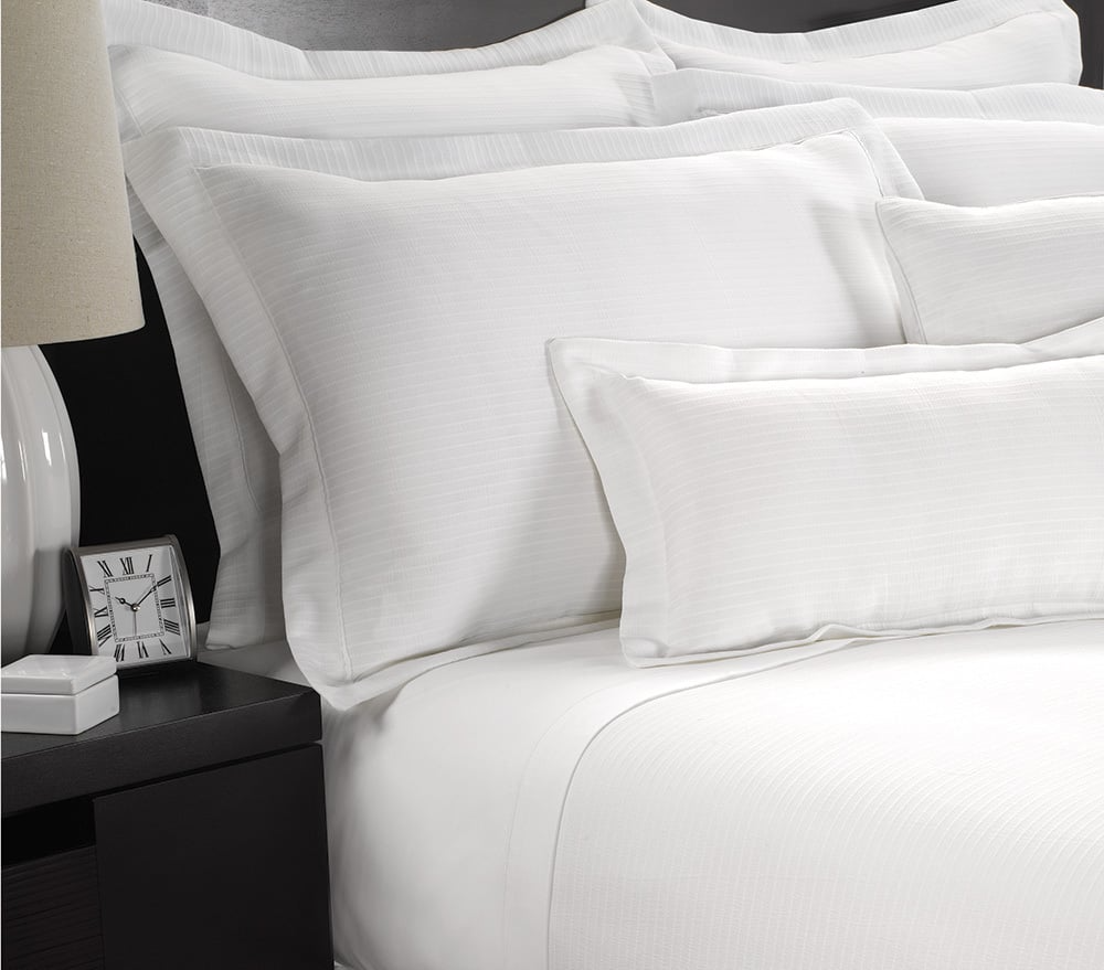 A neatly made bed features the Martex Wovens Decorative Top Sheet by WestPoint Home, with plush pillows, beside a nightstand that holds a white lamp and a small clock displaying 8:20.