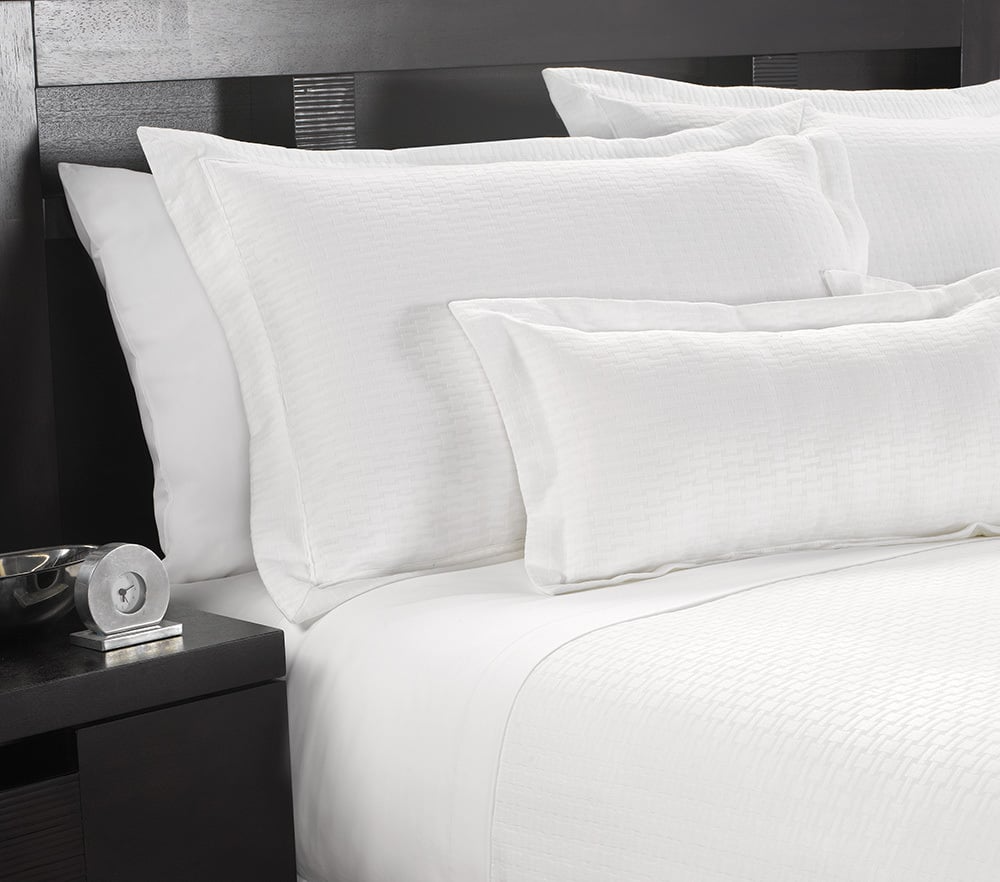 A neatly made bed features WestPoint Homes Martex Wovens Decorative Top Sheets and textured pillows, next to a dark wooden nightstand with a small silver clock and bowl.