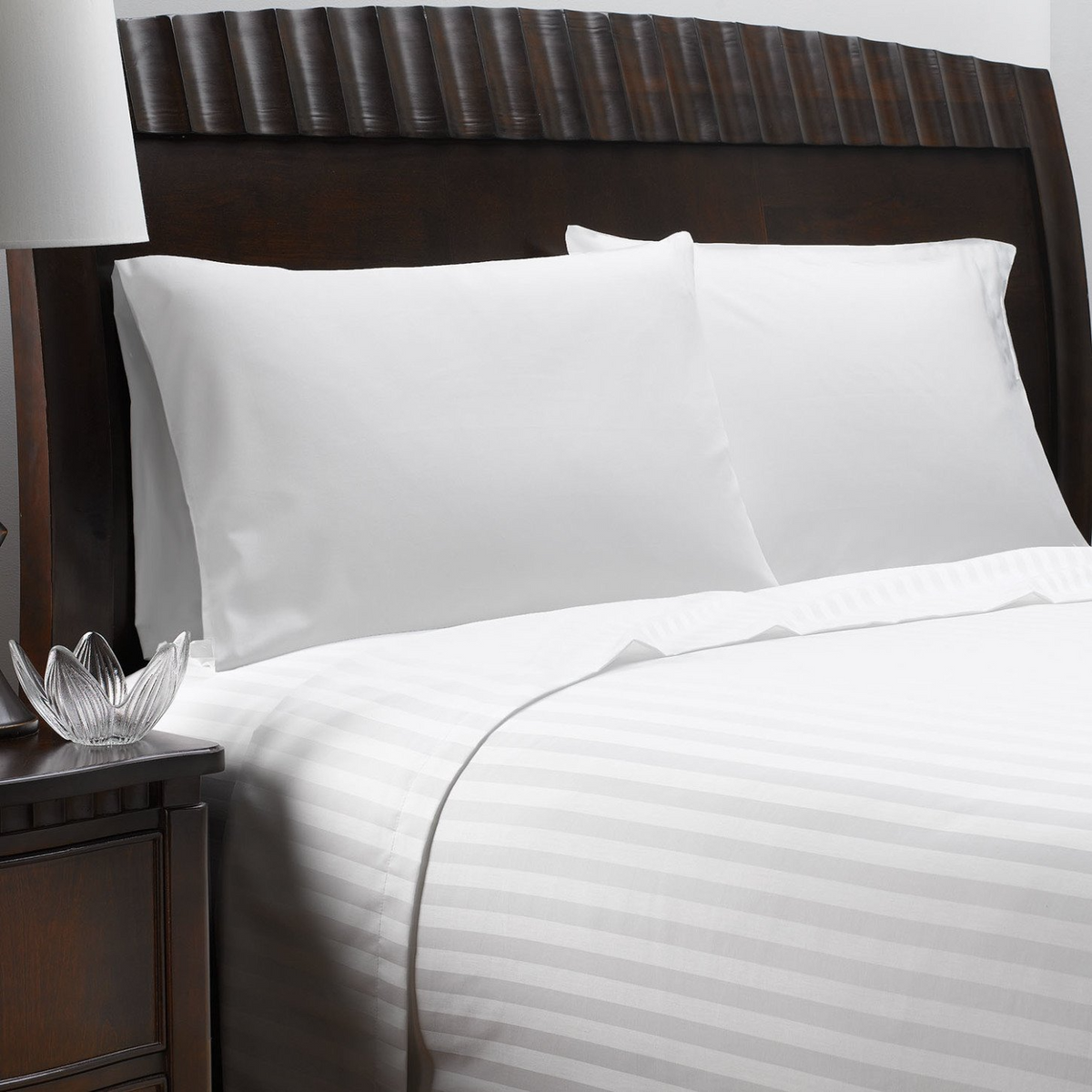A neatly made bed features the Martex Wovens Decorative Top Sheet by WestPoint Home in crisp white jacquard, with two pillows and a dark wooden headboard. A nightstand with a lamp and glass decor stands beside the bed.