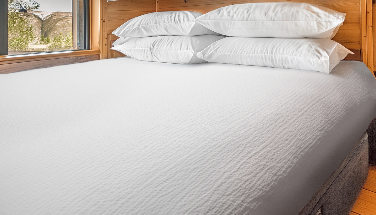 A neatly made bed features the Martex Wovens Decorative Top Sheet by WestPoint Home, accented with six stacked pillows in a wood-paneled room with a window showcasing a scenic outdoor view.