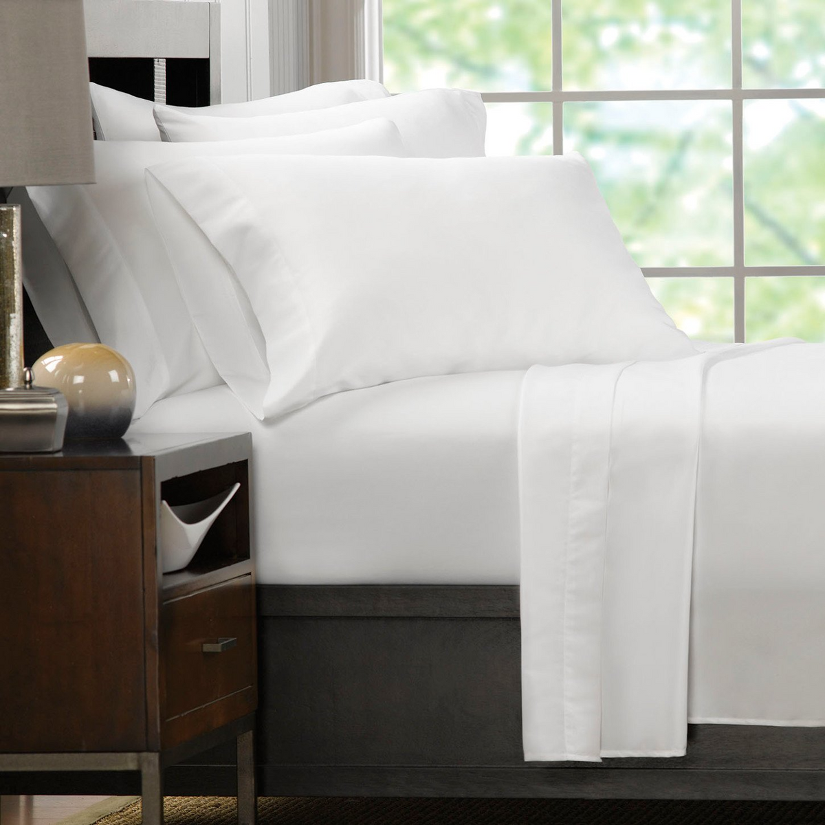 A bed made with WestPoint Homes Martex Ultra Touch Flat Sheet sits beside a wooden nightstand topped with a lamp and bowl, next to a large window revealing a softly blurred outdoor view.