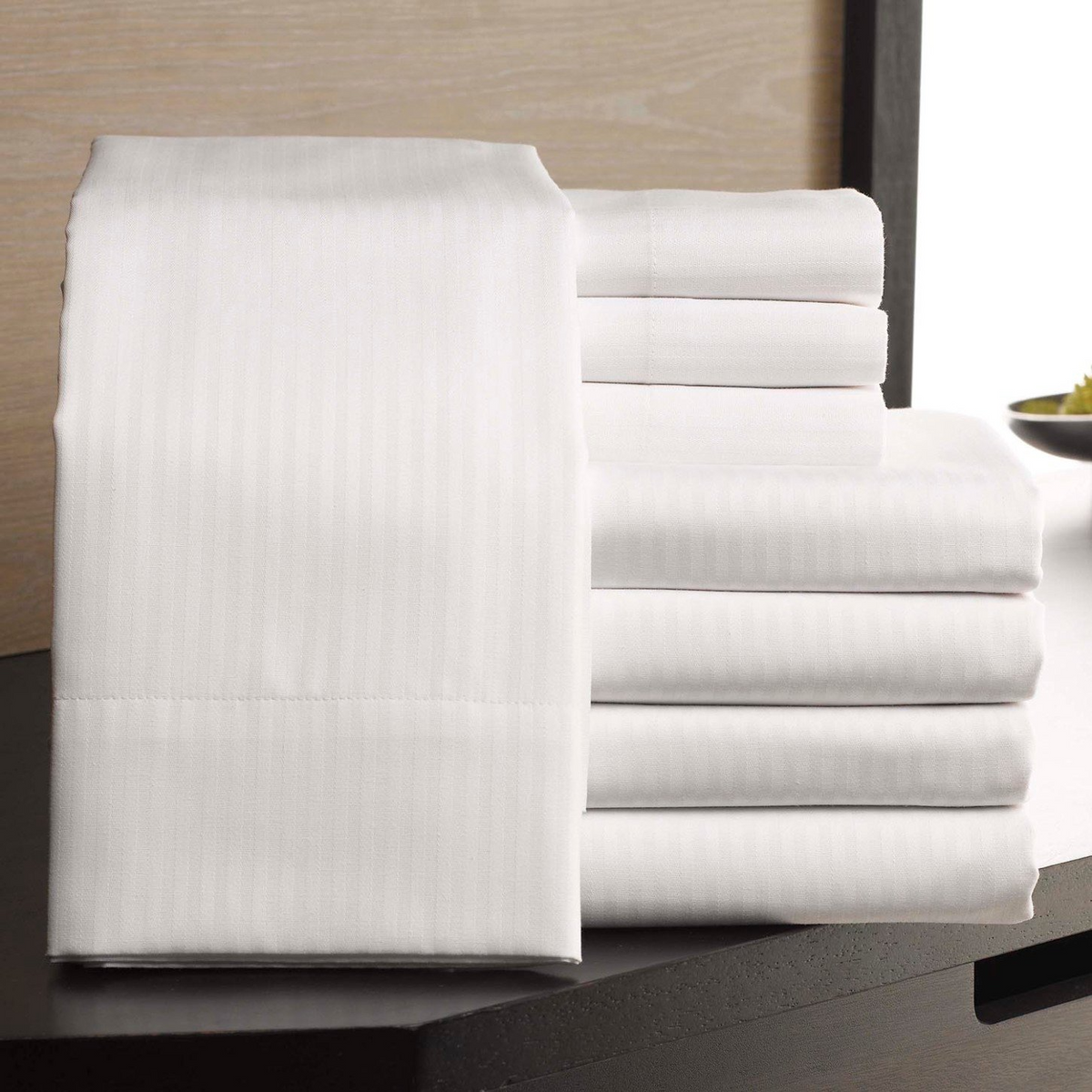 A neatly folded stack of WestPoint Home Patrician Tone on Tone Stripe fitted sheets is arranged on a dark surface, with one cotton-rich sheet draped to show its smooth, striped texture. The background has a light wood panel and a small decorative bowl.