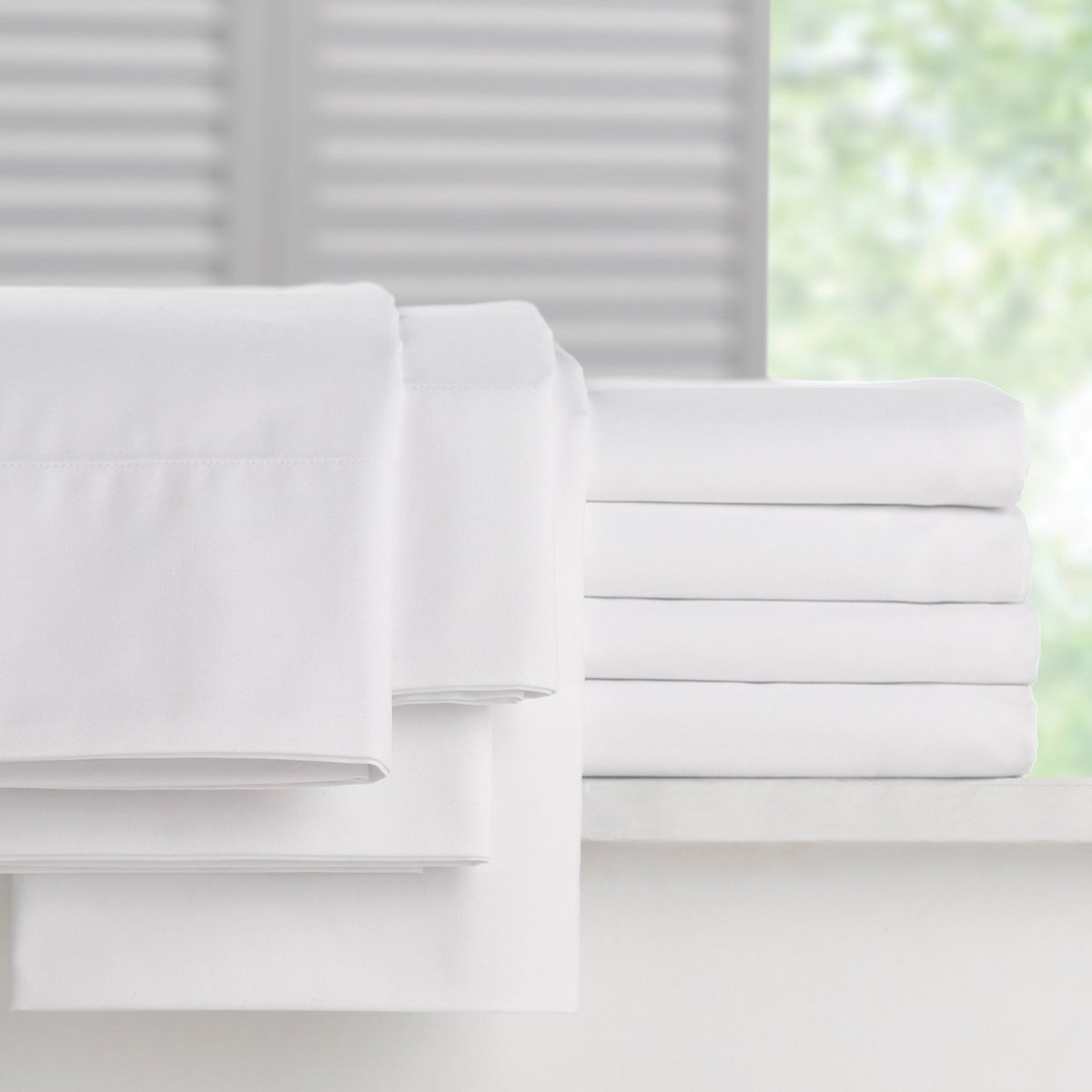 A neatly folded stack of WestPoint Home Martex Millennium 250 Bone sheets and pillow cases rests on a white surface, with a softly blurred background of a window and greenery outside.