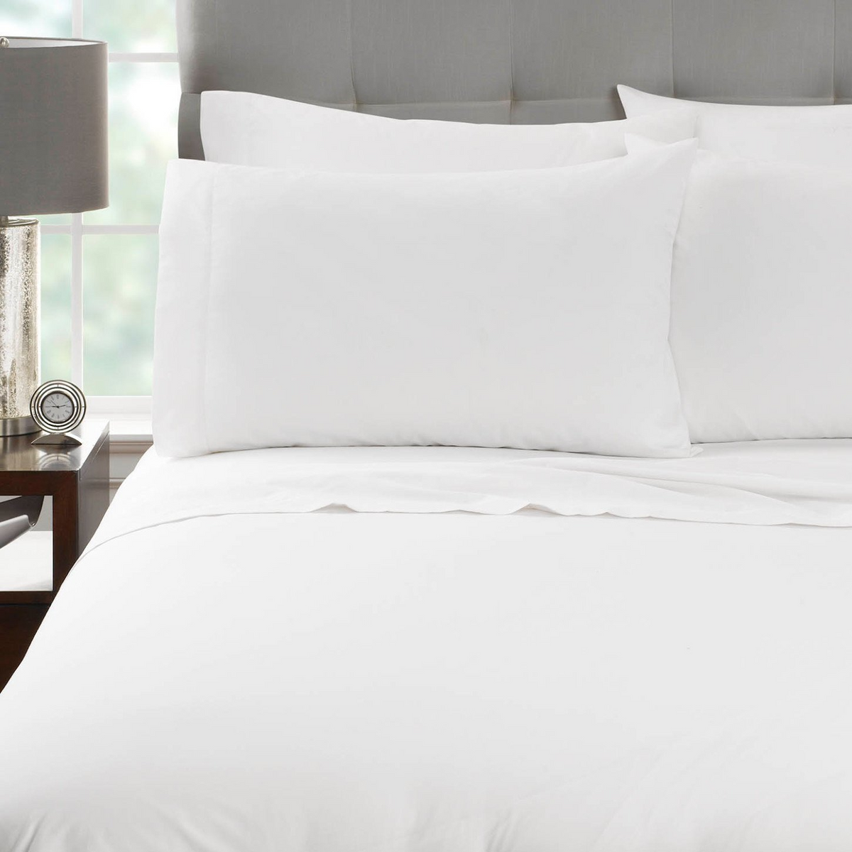 A neatly made bed features Martex Millennium 250 flat sheets by WestPoint Home, pillows, and a comforter beside a nightstand with a lamp and decor, set against a tufted gray headboard with a window in the background.