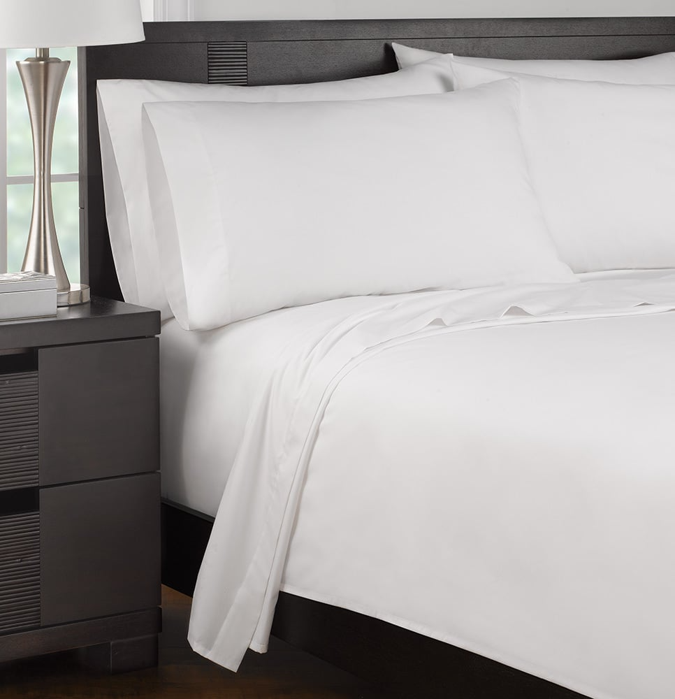 A neatly made bed with crisp white Martex DryFast 200 Flat Sheet by WestPoint Home sits next to a dark wooden nightstand featuring a silver lamp and a book.