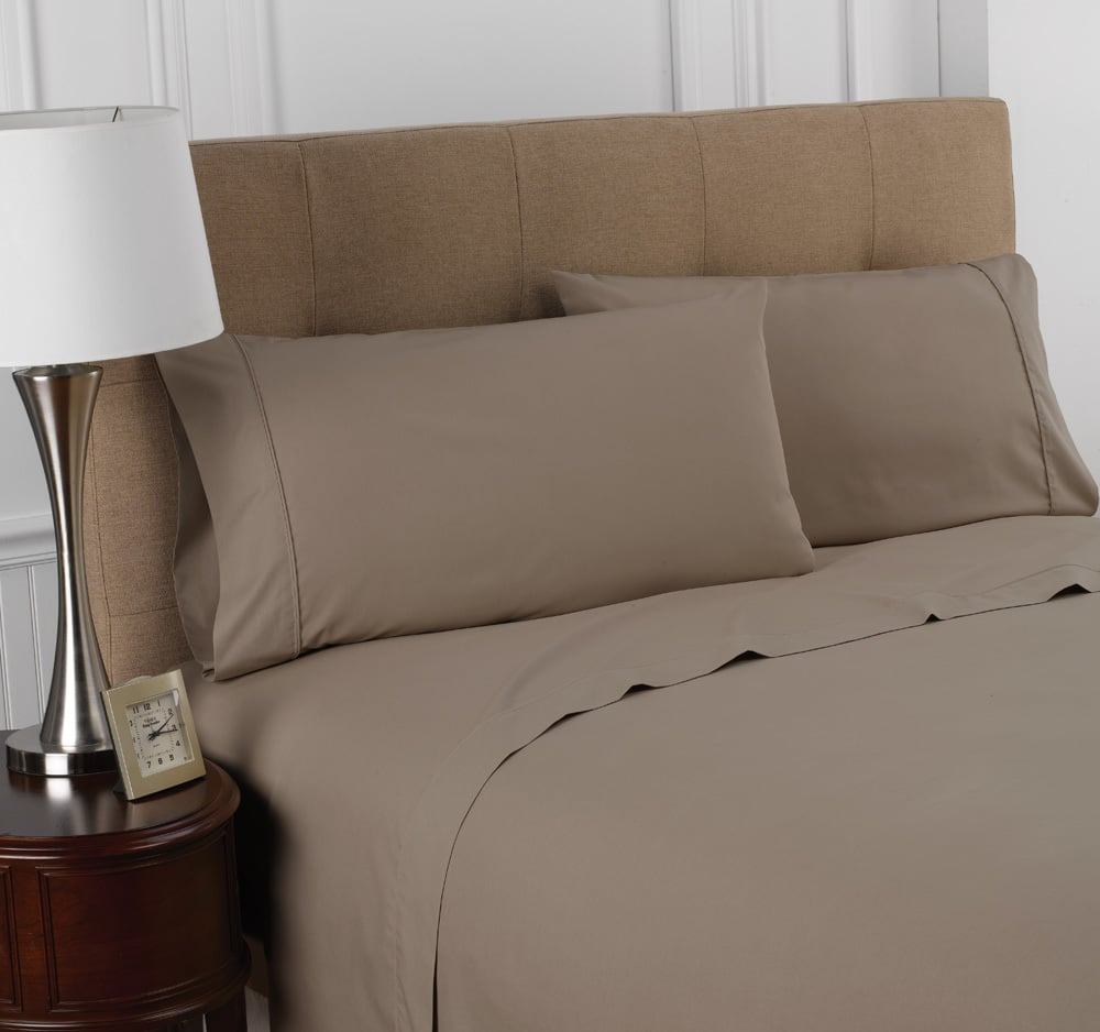 A bed neatly made with the Martex Colors 200 Flat Sheet by WestPoint Home in taupe, paired with cotton-polyester blend pillowcases, a beige upholstered headboard, wooden nightstand with clock, and modern silver lamp.