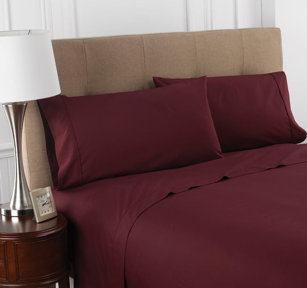 A neatly made bed with Martex Colors 200 pillowcases by WestPoint Home in burgundy, a beige upholstered headboard, a wooden nightstand with lamp, and a small framed photo against a white paneled wall.