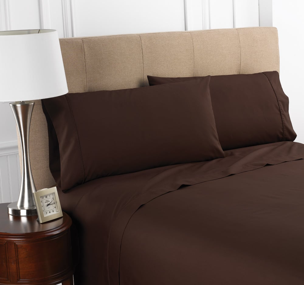 A neatly made bed with dark brown WestPoint Home Martex Colors 200 Flat Sheet and pillowcases, beige upholstered headboard, round wooden nightstand with lamp and clock, and white paneled walls in the background.