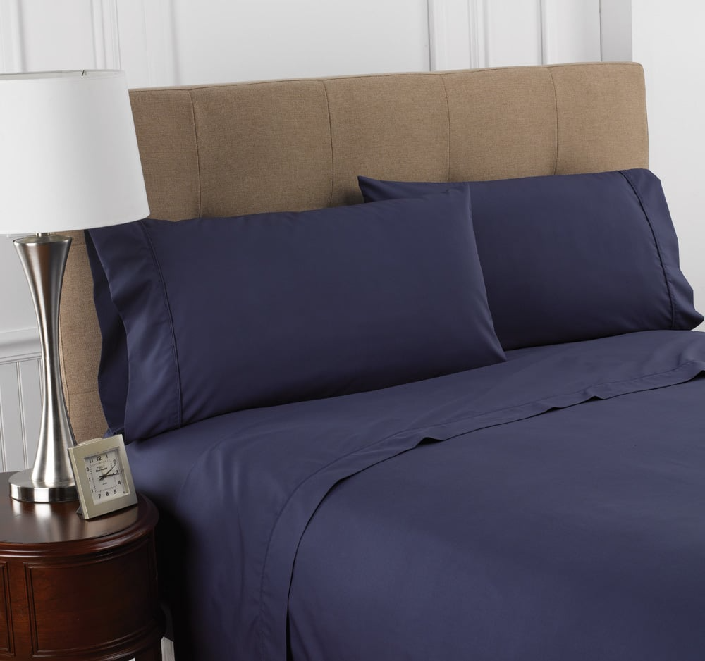 A neatly made bed with WestPoint Home Martex Colors 200 Flat Sheet in navy, beige upholstered headboard, and a round wooden nightstand holding a silver lamp and small clock.