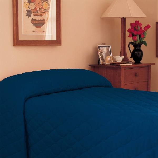 A bed with a dark blue Martex Mainspreads Solid bedspread by WestPoint Home is beside a wooden nightstand holding a black vase with red flowers, framed photos, and a beige lamp. A framed floral picture hangs on the beige wall above.