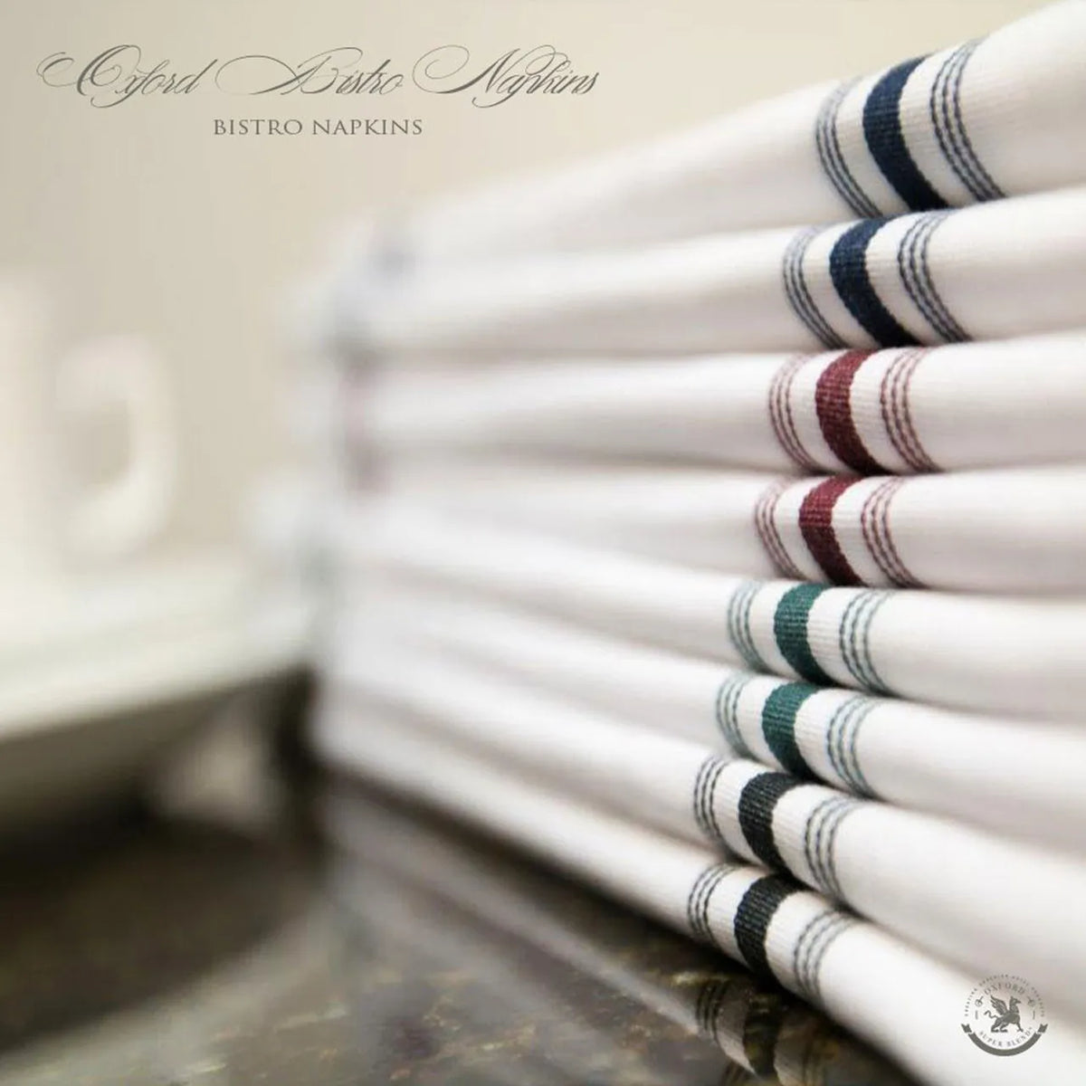 A stack of Ganesh Mills Bistro Napkins – White w/ Color Stripes (red, blue, green) sits on a countertop. The classic polyester napkins feature colorful striped edges and timeless bistro style.