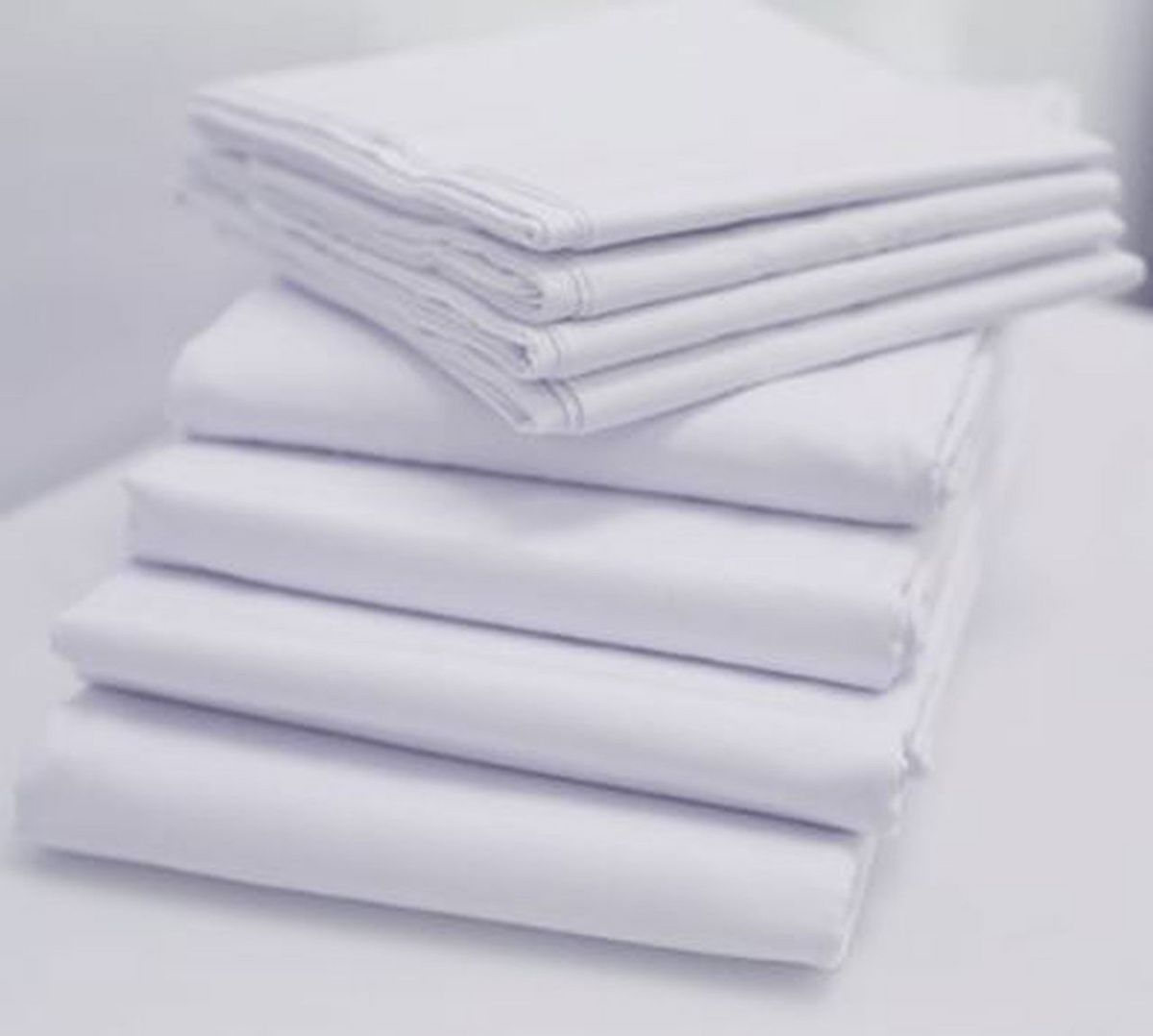 A neatly stacked pile of 1Concier Flat Sheet Solid Percale Blend bed sheets rests on a white surface, four on the bottom and three on top. A softly blurred background highlights their clean, orderly arrangement.