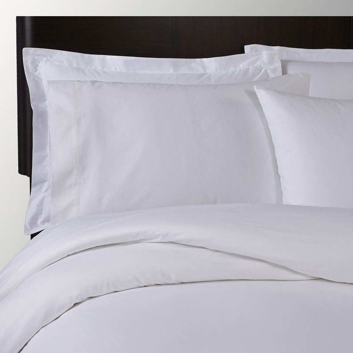 A neatly made bed with a crisp Duvet Cover Poly Percale T250 by 1Concier, two large white pillows with shams, and a white comforter against a dark wooden headboard.